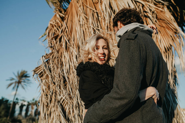Blonde woman smiling while she embraces at her boyfriend at sunset with a palm tree in the background. - Φωτογραφία, εικόνα