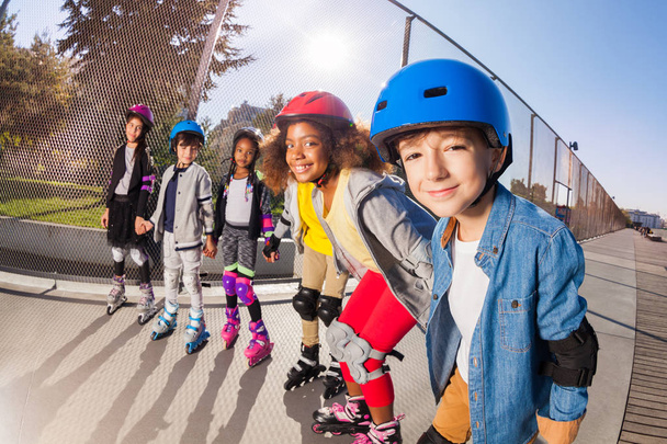 Big group of multiethnic boys and girls, roller skaters in safety helmets, holding hands standing in line at stadium outdoors - 写真・画像