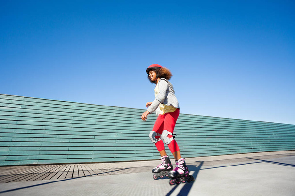 Side view portrait of African girl in helmet and protective gear, riding fast at rollerdrom against blue sky - Foto, immagini