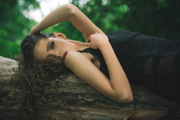 Woman with curly hairstyle in summer. Woman with long brunette hair relax on tree on nature. Hair care, therapy and treatment. Girl with makeup face, beauty. Fashion model with glamour look - Foto, Imagem