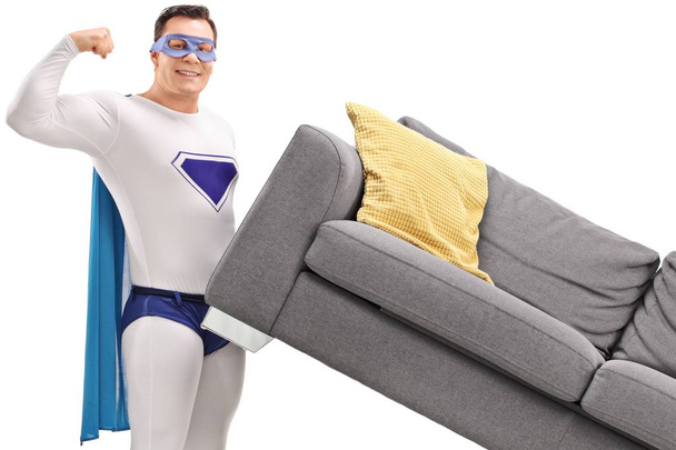 Young man in a superhero costume lifting a sofa and flexing his biceps isolated on white background - Фото, зображення