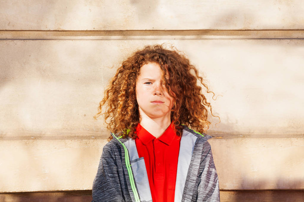 Portrait of teenage curly-haired boy standing against the wall outdoors and looking at camera - Foto, immagini