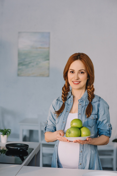 beautiful pregnant woman holding plate with ripe apples at kitchen - Valokuva, kuva