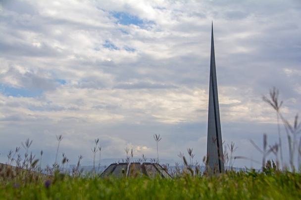 エレバン, アルメニアの劇的な曇り空の下の濡れた芝生を背景にアルメニアの集団虐殺の大虐殺のモニュメント記念碑。低角度のビュー。ぼやけ前景 - 写真・画像