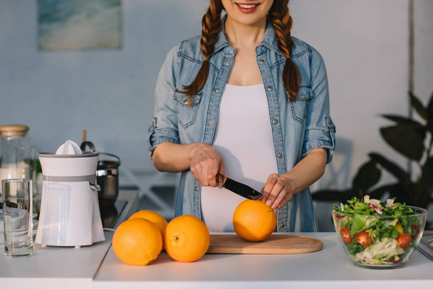 cropped image of smiling pregnant woman cutting oranges at kitchen - Fotó, kép