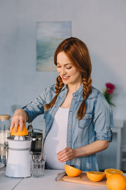 attractive pregnant woman preparing homemade orange juice at kitchen - Photo, Image