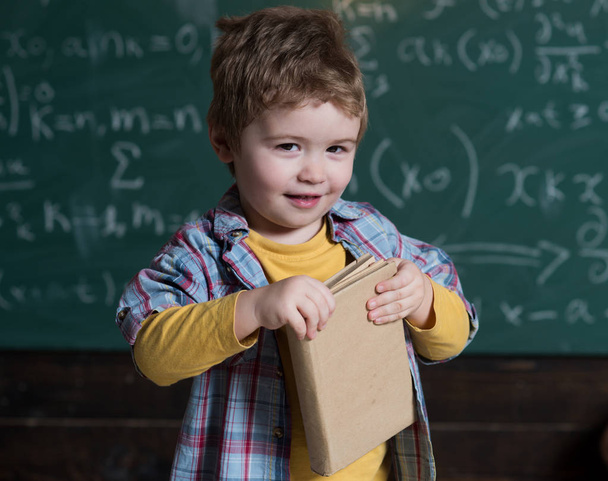 Smart child on smiling face, holds book in hands. Kid, preschooler or first former, chalkboard on background, defocused. Studying concept. Boy looks cute with book, kid likes to study. - Photo, Image