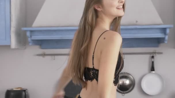 Young beautiful sexy Caucasian girl in black lingerie walking around the kitchen, smiling, looking at the camera, temptation, sexy female body, belly, butt, white sunlight in the background. 60 fps - Video