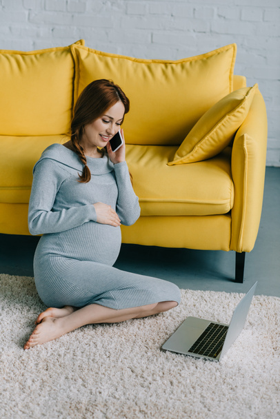 attractive pregnant woman sitting on floor and talking by smartphone in living room - Фото, зображення