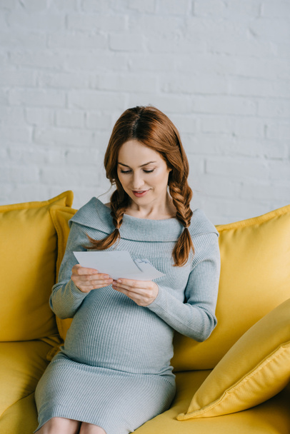 attraktive schwangere Frau sitzt auf dem Sofa und betrachtet Fotos im Wohnzimmer - Foto, Bild