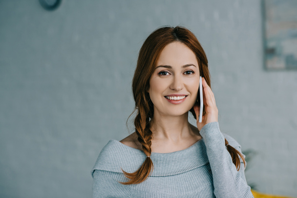 attractive smiling woman talking by smartphone and looking at camera at home - Fotografie, Obrázek
