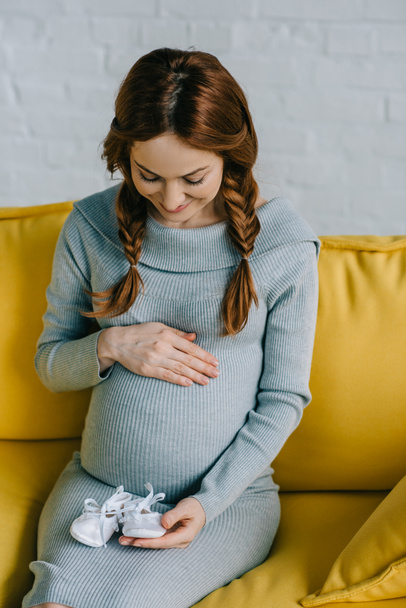 atractiva mujer embarazada tocando el abdomen y mirando los zapatos recién nacidos en la sala de estar
 - Foto, imagen