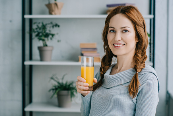 Attractive smiling woman holding glass of orange juice and looking at camera at home - 写真・画像