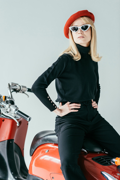 Fashionable blonde girl in red beret posing by vintage scooter isolated on grey - Photo, Image
