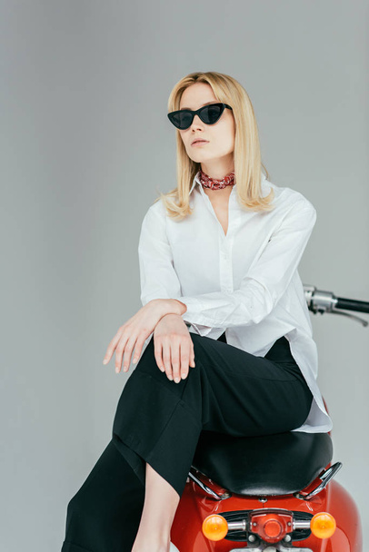 Stylish pretty woman in sunglasses posing on red scooter isolated on grey - Zdjęcie, obraz