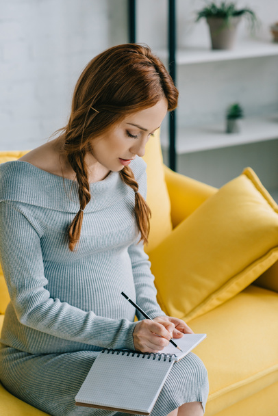 attractive pregnant woman writing something to notebook in living room - Foto, Imagen