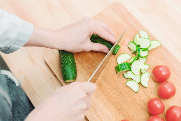 close-up of female hands cut into fresh cut cucumbers on a wooden cutting board next to pink tomatoes. The concept of homemade vegetarian cuisine and healthy eating and lifestyle - Foto, Bild
