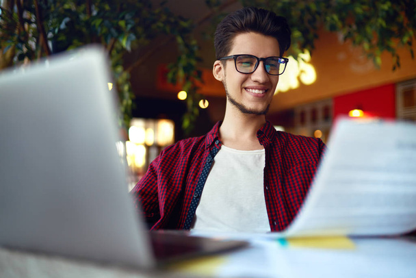 Smiling young hipster man in glasses with laptop on the table. Programmer, web developer, designer working at office comparing mobile and desktop website versions. - Foto, Imagem