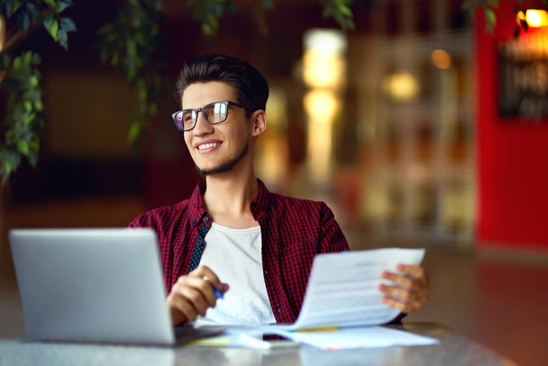 Um jovem hipster sorridente de óculos com laptop na mesa. Programador, desenvolvedor web, designer trabalhando no escritório comparando versões de sites móveis e desktop
. - Foto, Imagem