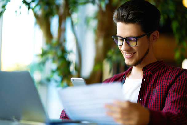 Glimlachende man van de jonge hipster in glazen met laptop op tafel. Programmeur, webontwikkelaar, ontwerper werkt op kantoor mobiele en desktop website versies vergelijken. - Foto, afbeelding