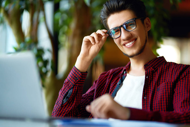 Glimlachende man van de jonge hipster in glazen met laptop op tafel. Programmeur, webontwikkelaar, ontwerper werkt op kantoor mobiele en desktop website versies vergelijken. - Foto, afbeelding