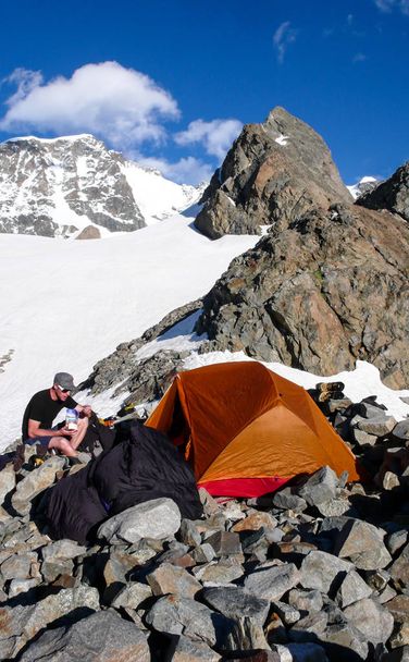 чоловічий гірський альпініст їсть у базовому таборі біля помаранчевого намету з вражаючим гірським пейзажем навколо нього
 - Фото, зображення