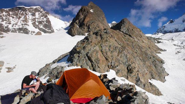 alpiniste mâle mangeant au camp de base près d'une tente orange avec un paysage de montagne spectaculaire autour de lui
 - Photo, image