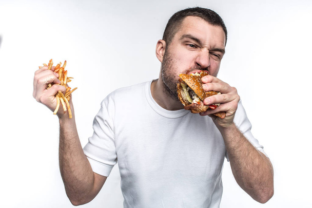 This guy is very delight of junk food. He is biting a big piece of burger and holding a full hand of french fries covered with ketchup. Young amn likes to eat oily meal. Isolated on white background. - Fotó, kép