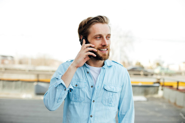 casual young man in blue shirt having a conversation, talking on cellphone, somewhere outside, smiling and enjoying this conversation - Photo, image