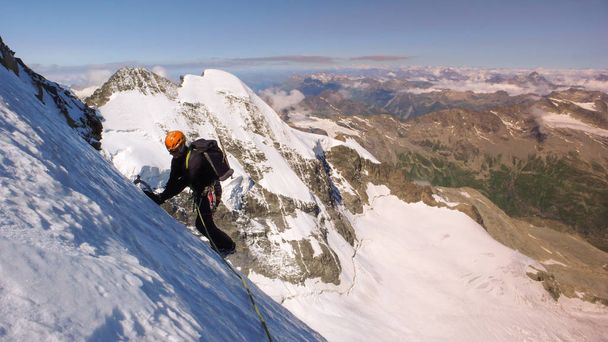 miespuolinen vuorikiipeilijä korkealla alppijäätiköllä upealla näköalalla upealle vuoristomaisemalle hänen takanaan
 - Valokuva, kuva