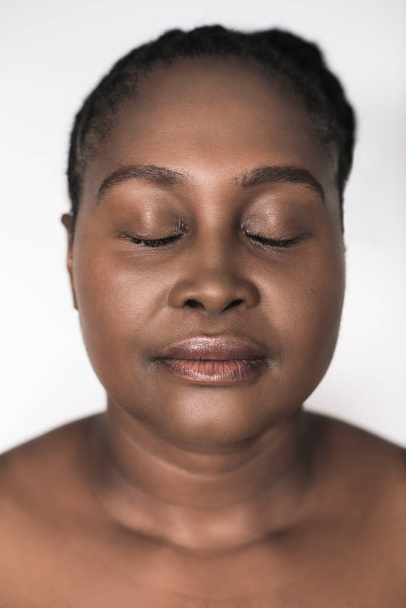 Nahaufnahme einer jungen afrikanischen Plus-Size-Frau mit perfektem Teint, die mit geschlossenen Augen vor weißem Hintergrund steht - Foto, Bild