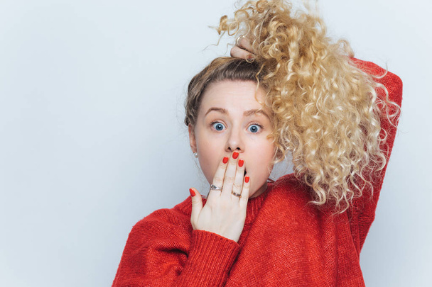 Bugged eyed emotional funny blonde female with pony tail and curly hair, covers mouth with hand, doesn`t expect to hear shocking news from friend, isolated over white background. Omg concept - Foto, imagen
