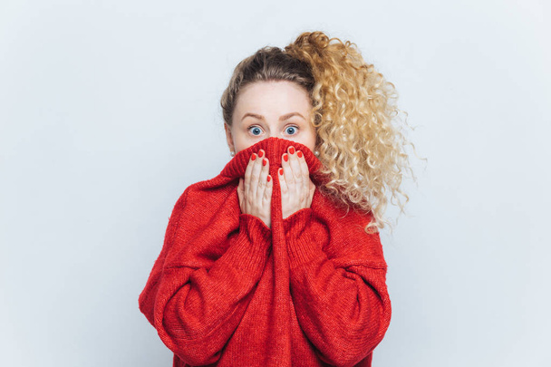 Vyděšená žena omráčena vidí její fobie, pokrývá tvář s límcem červený svetr, zírá na fotoaparát s vyděšený pohled, izolované nad bílým studio pozadí. Koncept lidí, reakce a emoce - Fotografie, Obrázek