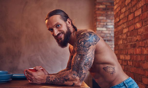 Un hombre guapo barbudo tatuado con un corte de pelo elegante y cuerpo musculoso, bebe café, apoyado en una mesa
. - Foto, Imagen