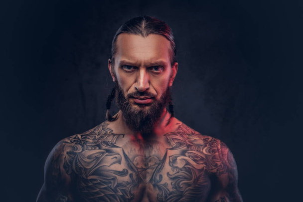 Retrato de cerca de un macho musculoso barbudo tatuado con un corte de pelo elegante, aislado sobre un fondo oscuro
. - Foto, Imagen