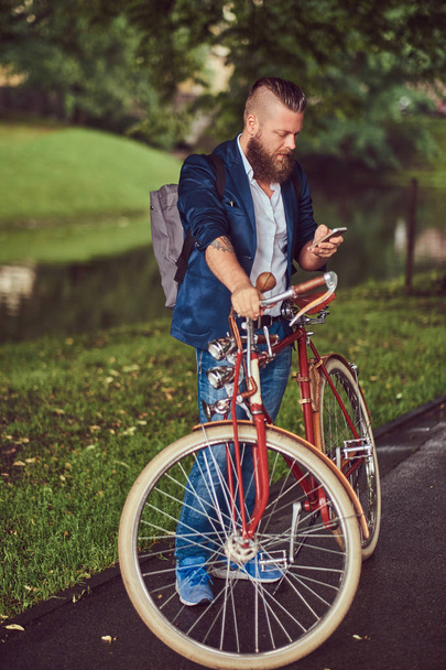 Ταξιδιώτης ντυμένος με casual ρούχα με ένα σακίδιο, χρησιμοποιώντας ένα smartphone, χαλαρώνοντας σε ένα πάρκο της πόλης μετά την ιππασία σε ένα ρετρό ποδήλατο. - Φωτογραφία, εικόνα