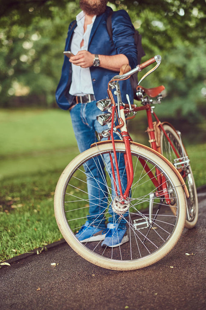 Matkustaja pukeutunut rento vaatteita ja aurinkolasit reppu, älypuhelimella, rentouttava kaupungin puistossa ajettuaan retro polkupyörällä
. - Valokuva, kuva
