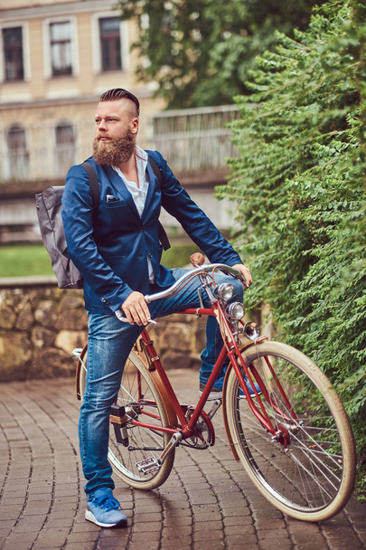 Ένας γενειοφόρος άνδρας με ένα μοντέρνο κούρεμα ντυμένοι με πρόχειρα ρούχα με ένα σακίδιο, κάθεται σε ένα ρετρό ποδήλατο σε ένα πάρκο της πόλης. - Φωτογραφία, εικόνα