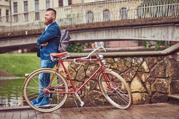 Parrakas mies, jolla on tyylikäs kampaus, joka on pukeutunut rentoihin vaatteisiin reppuineen, seisoo retro-polkupyörän kanssa joen lähellä kaupungin puistossa
. - Valokuva, kuva