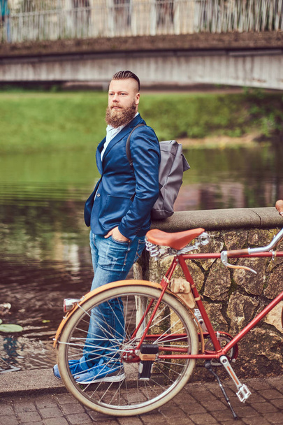 Γενειοφόρος άνδρας με ένα μοντέρνο κούρεμα ντυμένοι με πρόχειρα ρούχα με ένα σακίδιο, στέκεται με ένα ρετρό ποδήλατο κοντά σε ένα πάρκο της πόλης στον ποταμό. - Φωτογραφία, εικόνα