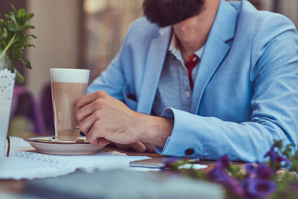 Πορτρέτο ενός μόδας γενειοφόρου άνδρα με ένα μοντέρνο κούρεμα, κατέχει ένα ποτήρι καπουτσίνο, κάθεται σε μια υπαίθρια καφετέρια. - Φωτογραφία, εικόνα