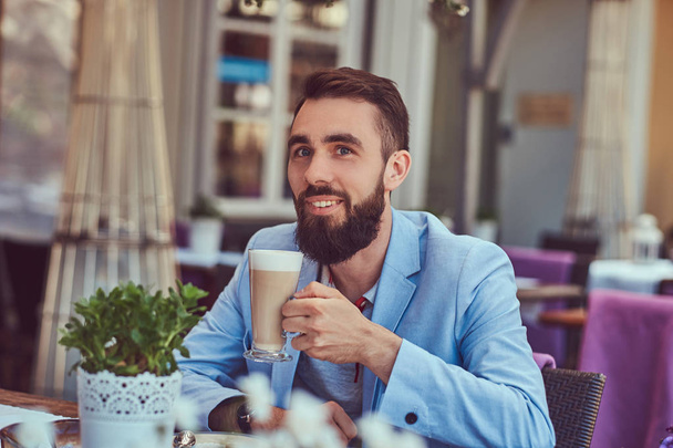 Portret van een modieuze bebaarde man met een stijlvol kapsel, drinkt een glas voor een cappuccino, zittend in een café in de buitenlucht. - Foto, afbeelding