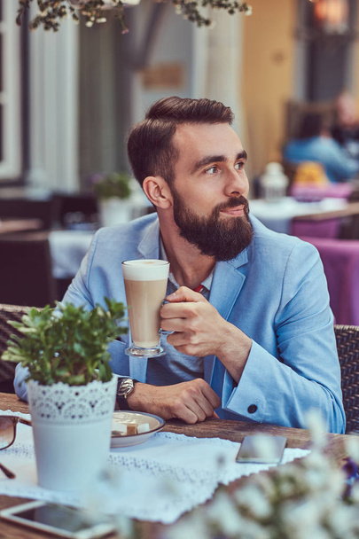 Portrait of a fashionable bearded male with a stylish haircut, drinks a glass of a cappuccino, sitting in a cafe outdoors. - Foto, Bild