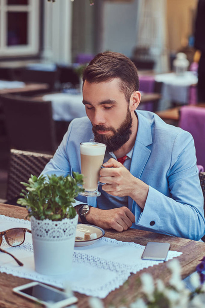 Retrato de un hombre barbudo de moda con un corte de pelo elegante, bebe un vaso de un capuchino, sentado en un café al aire libre
. - Foto, Imagen