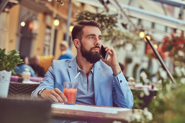 Telefonla konuşan bir moda sakallı işadamı şık bir saç kesimi ile portresi bir kafe mekanlarda oturan bir suyu bir bardak içer. - Fotoğraf, Görsel
