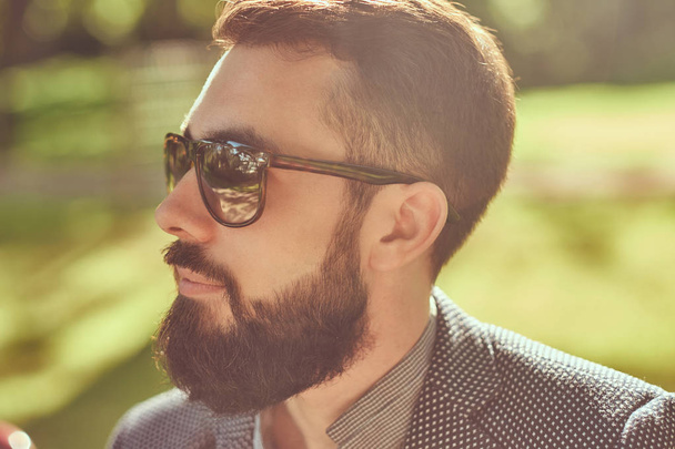 Ritratto ravvicinato di un maschio barbuto con un taglio di capelli elegante, in una giacca e occhiali da sole, seduto su una panchina in un parco
. - Foto, immagini