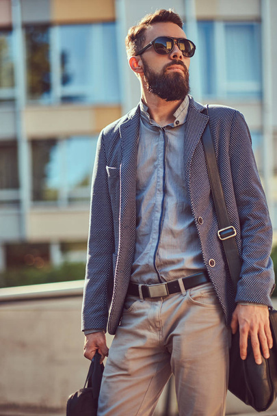 Retrato de um trabalhador barbudo vestindo roupas casuais e óculos de sol, com uma bolsa, em pé em uma rua da cidade contra um arranha-céu
. - Foto, Imagem