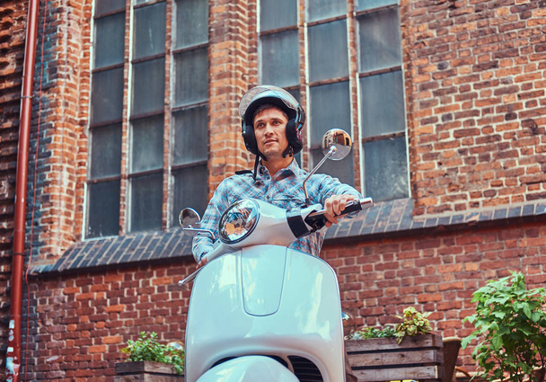 Bello uomo che indossa abiti casual in casco, cavalcando su uno scooter classico retrò, lungo le vecchie strade in un'Europa
. - Foto, immagini