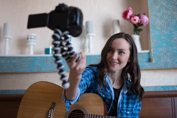 Молодая девушка с акустической гитарой в спальне записывает блок на DSLR камеру, держа его перед собой на gorillapod
 - Фото, изображение