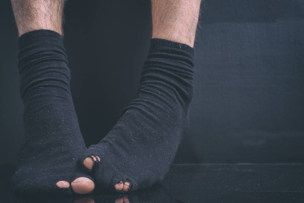 Τα πόδια του η κακή του οφειλέτη σε μαύρες κάλτσες holey σε μαύρο φόντο - Φωτογραφία, εικόνα